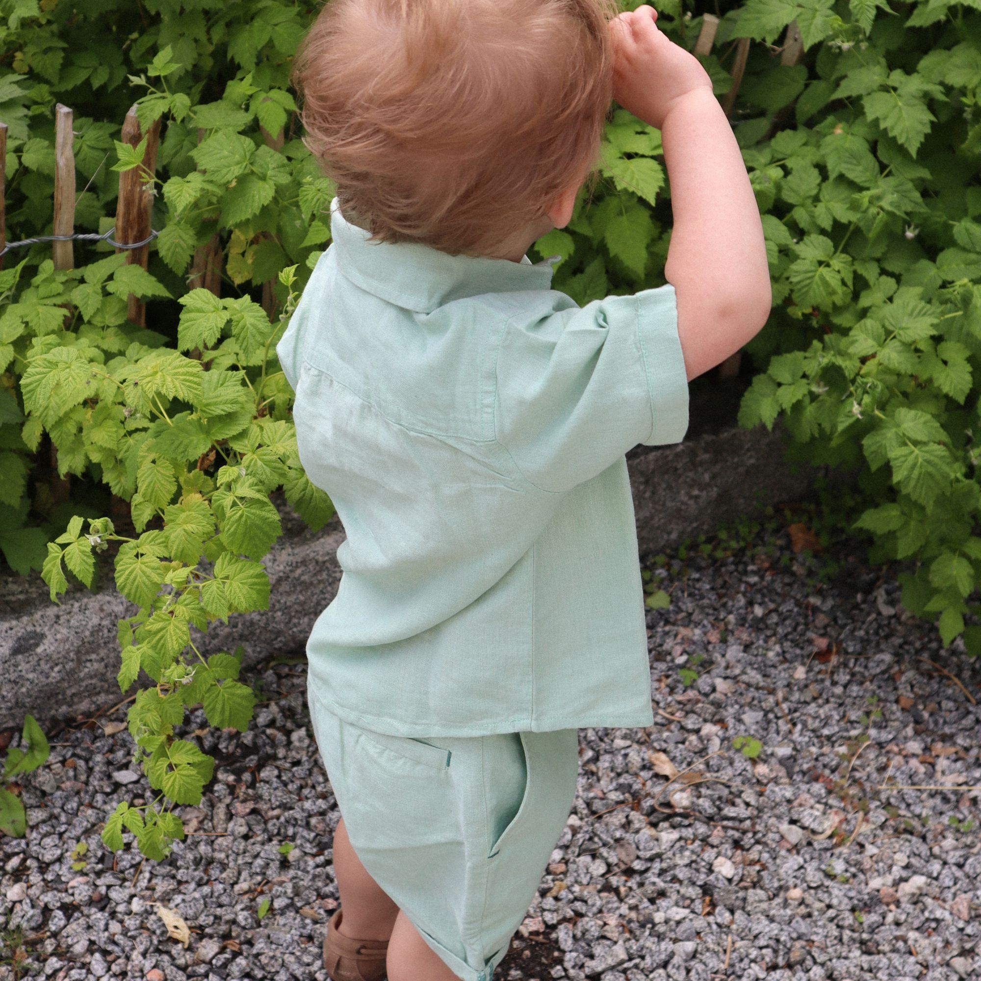 Mintgrønn kortermet linskjorte og matchende shorts på et barn i en frodig hage fra Geggamoja.