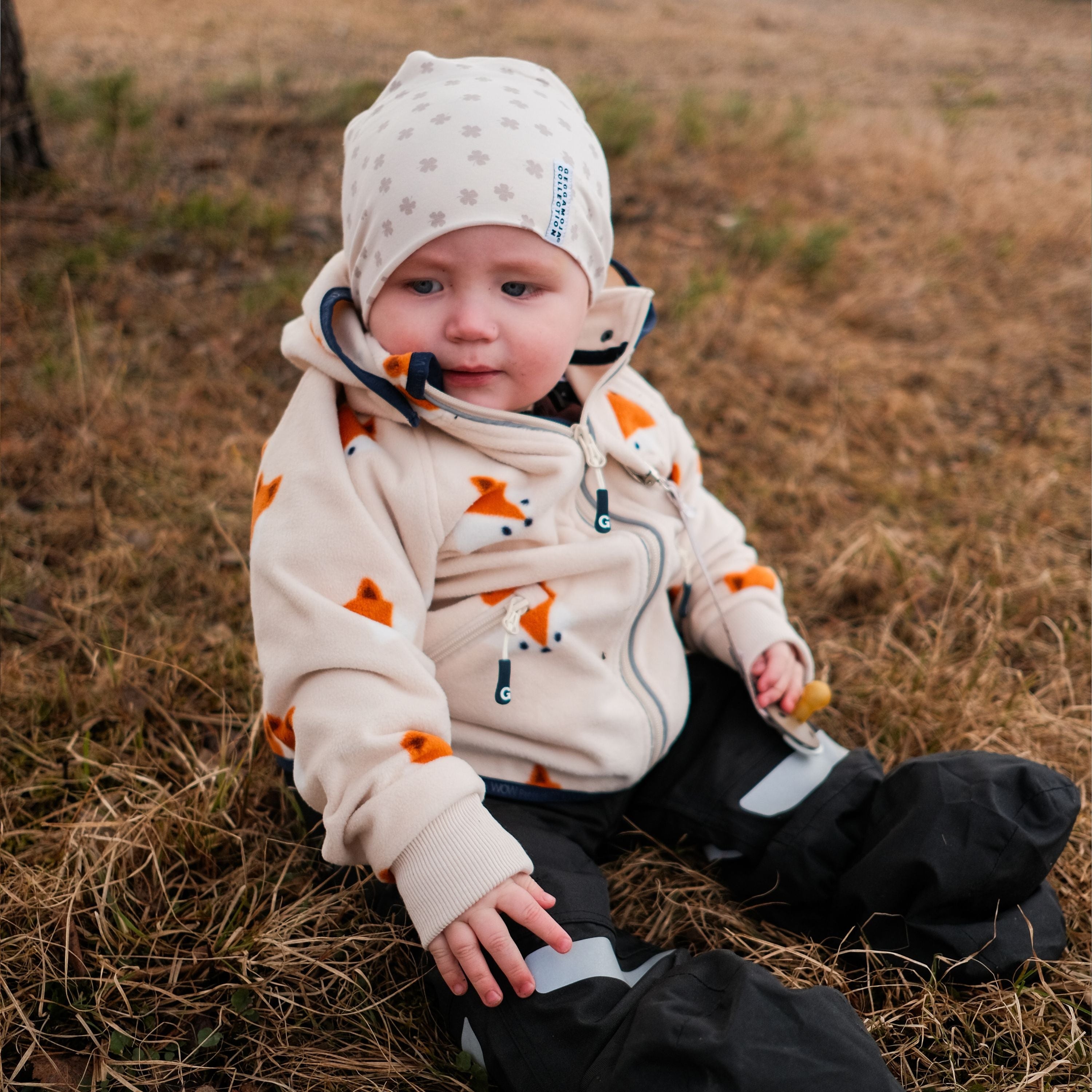 Beige mössa i bambu med klövermönster för baby från Geggamoja på en sittande bebis.